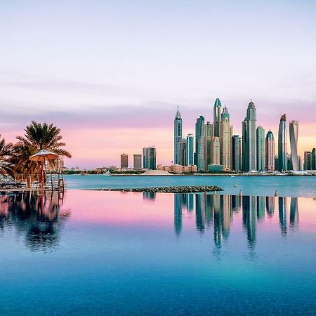 Dukes The Palm, A Royal Hideaway Hotel Dubai Esterno foto The image showcases a stunning skyline view of a modern city, likely during sunrise or sunset, as indicated by the beautiful pastel colors in the sky. The foreground features a calm body of water, reflecting the skyscrapers and the colorful sky. Lush