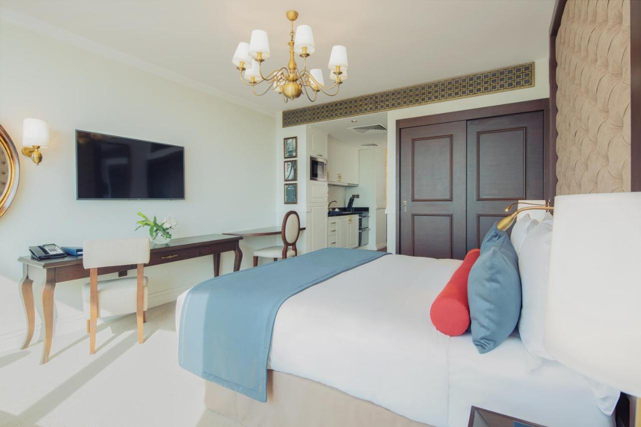 Dukes The Palm, A Royal Hideaway Hotel Dubai Esterno foto The photo depicts a modern hotel room featuring a neatly made queen-sized bed with decorative pillows in shades of blue and red. A stylish chandelier hangs from the ceiling, providing warm lighting. Opposite the bed, there is a wall-mounted flat-scre