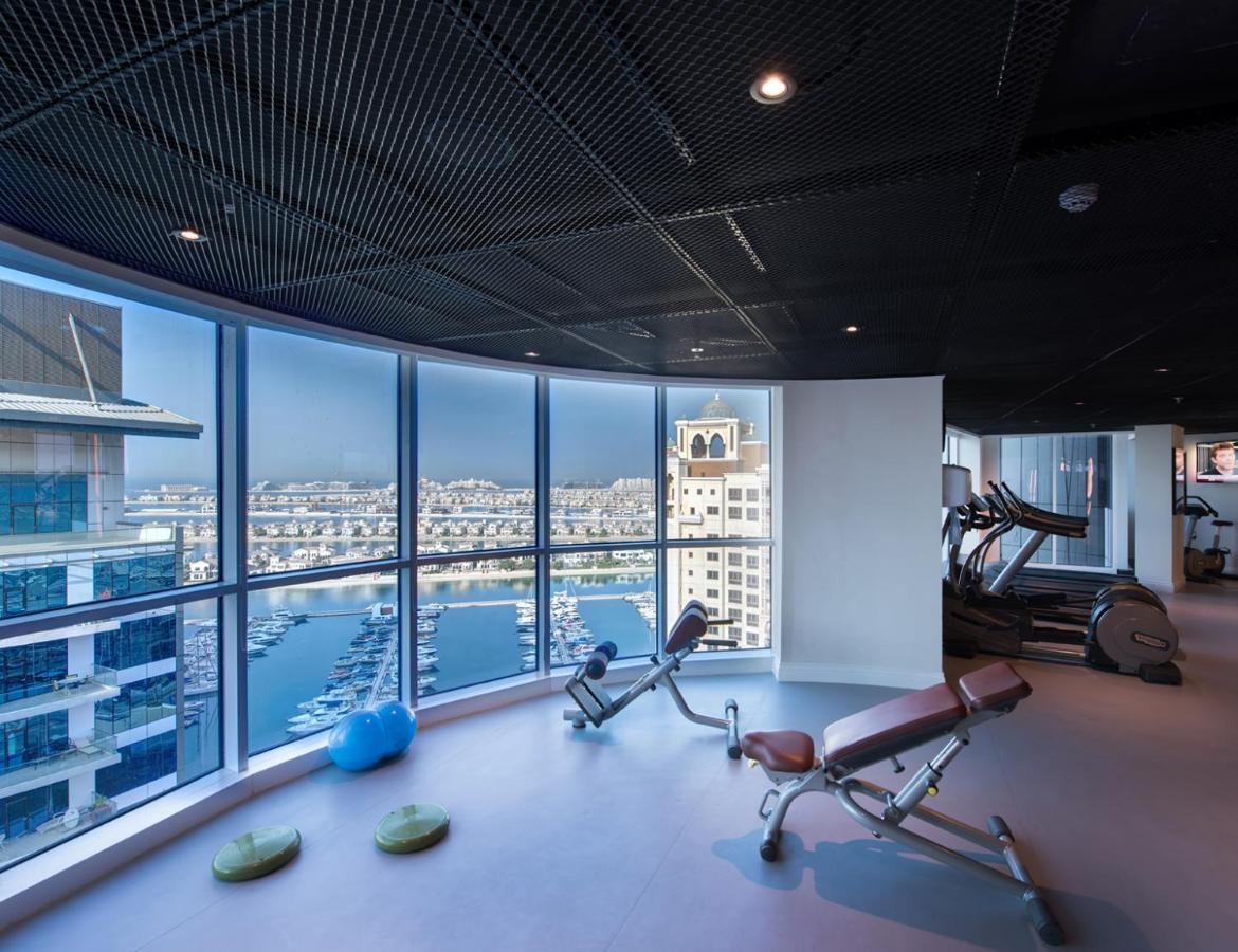 Dukes The Palm, A Royal Hideaway Hotel Dubai Esterno foto The photo shows a modern gym with large windows offering a panoramic view of a marina or waterfront area. Inside, there are exercise machines, including a couple of benches for weight training. The flooring is smooth, and there are a few exercise acc