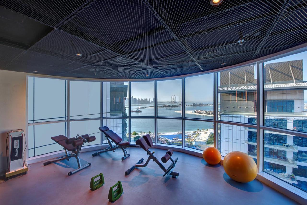 Dukes The Palm, A Royal Hideaway Hotel Dubai Esterno foto The photo shows a modern gym or fitness area that features large windows with a view of a waterfront and skyline. Inside, there are several pieces of fitness equipment, including adjustable benches and exercise balls. The design is sleek and contempo