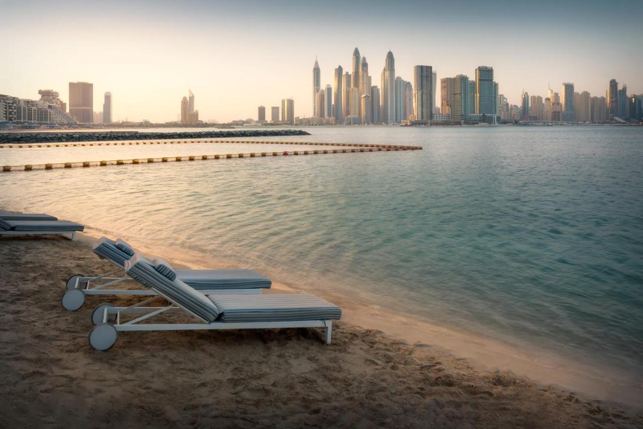 Dukes The Palm, A Royal Hideaway Hotel Dubai Esterno foto The photo shows a tranquil beach scene with several lounge chairs lined up on the sand, facing a calm body of water. In the background, a modern skyline is visible, featuring tall buildings and skyscrapers. The atmosphere appears serene, possibly dur