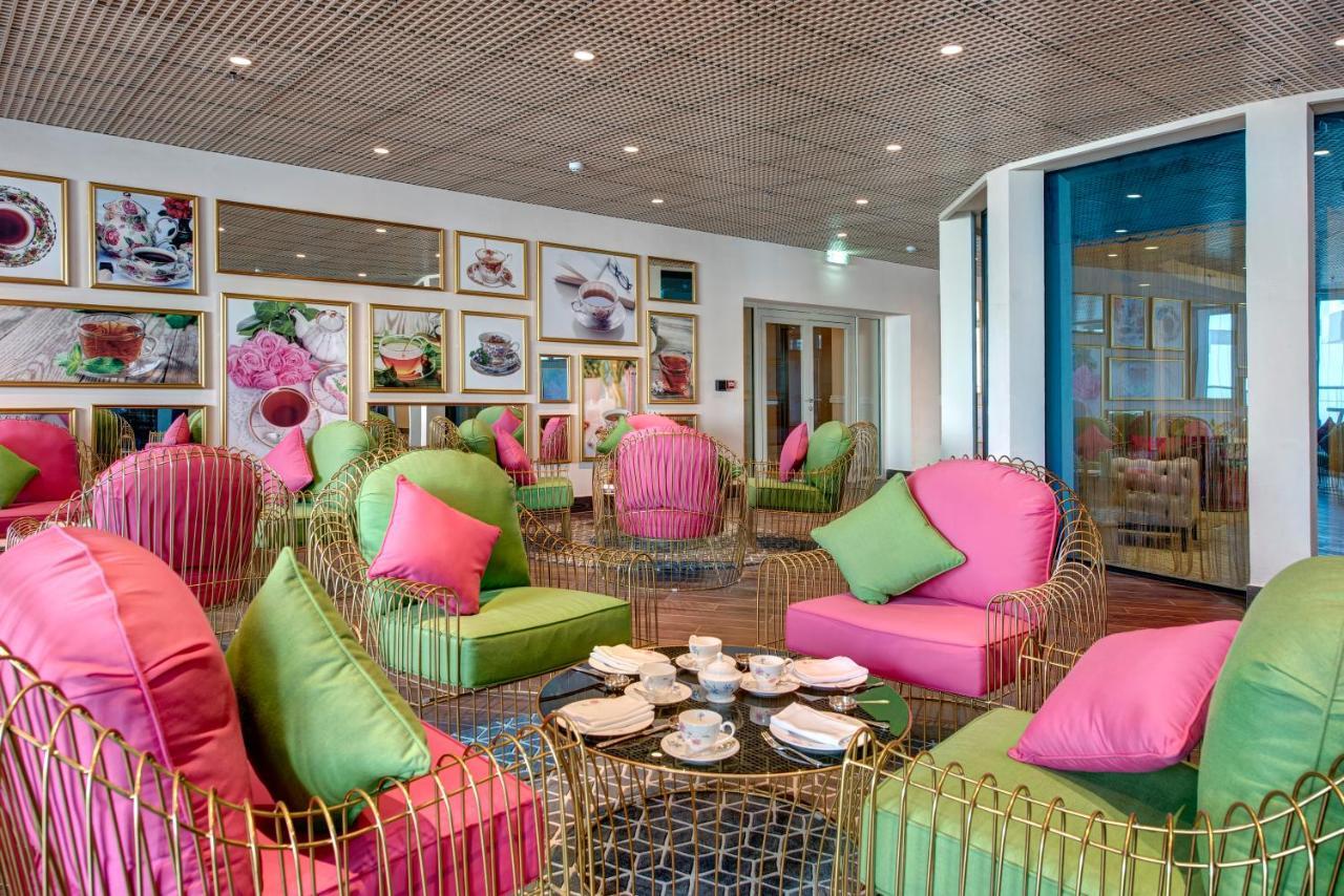 Dukes The Palm, A Royal Hideaway Hotel Dubai Esterno foto The photo shows a stylish and modern interior space, possibly a cafe or lounge. The seating area features elegant chairs with gold frames and vibrant cushions in shades of pink and green. There are several round tables with tea sets and small plates 