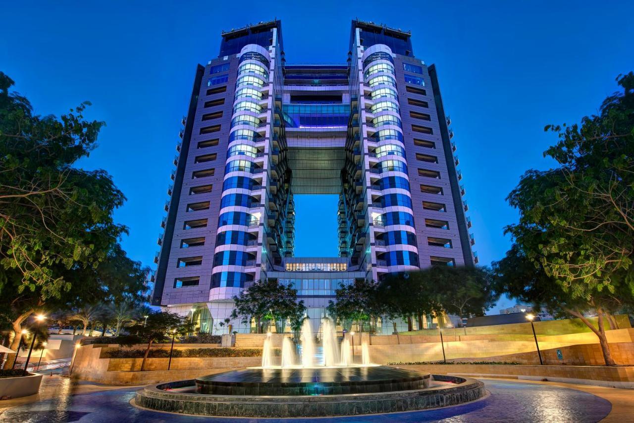 Dukes The Palm, A Royal Hideaway Hotel Dubai Esterno foto The photo shows a modern, tall building with a distinctive architectural design, featuring multiple floors and large windows. The structure is illuminated at dusk, highlighting its design and colors. In the foreground, there is a circular fountain wi