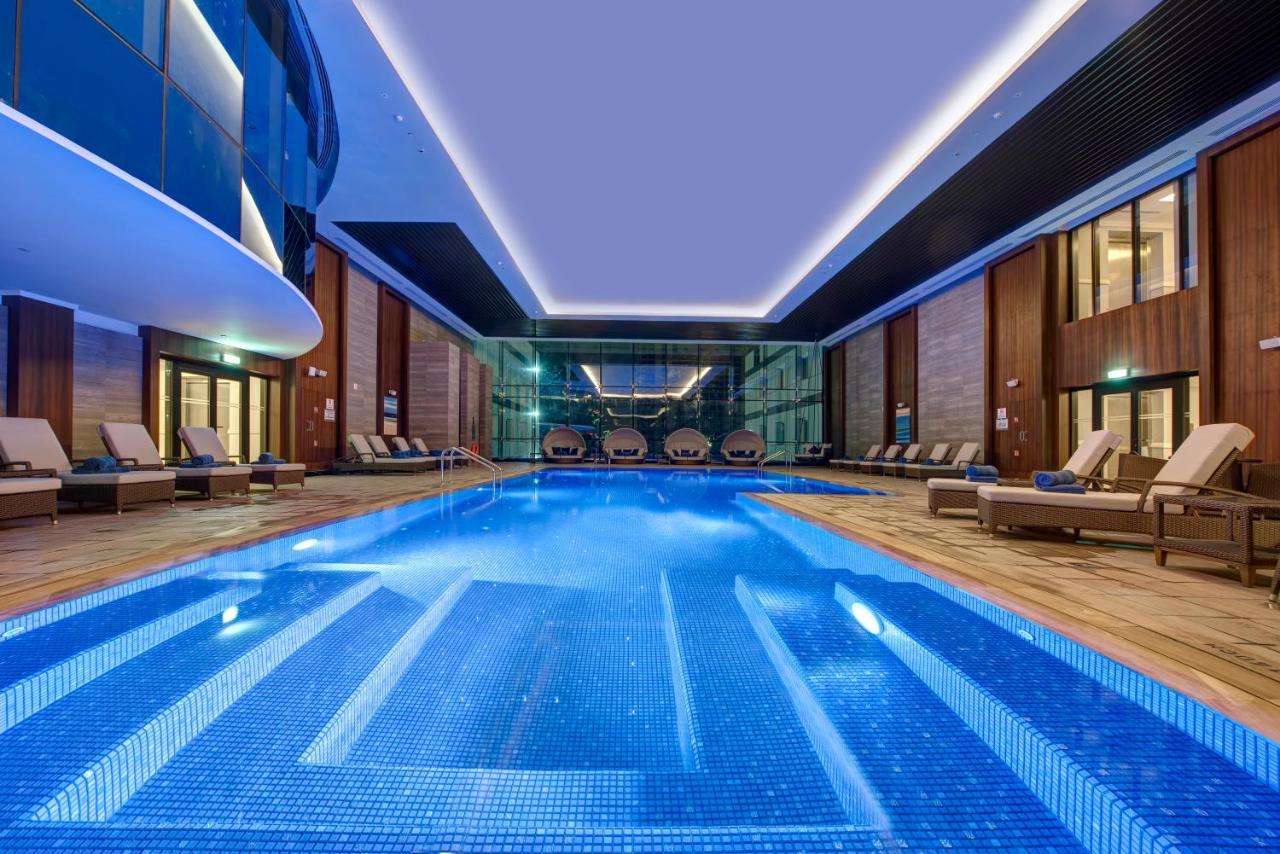 Dukes The Palm, A Royal Hideaway Hotel Dubai Esterno foto The photo shows a luxurious indoor swimming pool area. The pool is rectangular and features a blue tiled bottom, creating a vibrant hue in the water. Surrounding the pool are comfortable lounge chairs lined up along the edges. The area has large glas