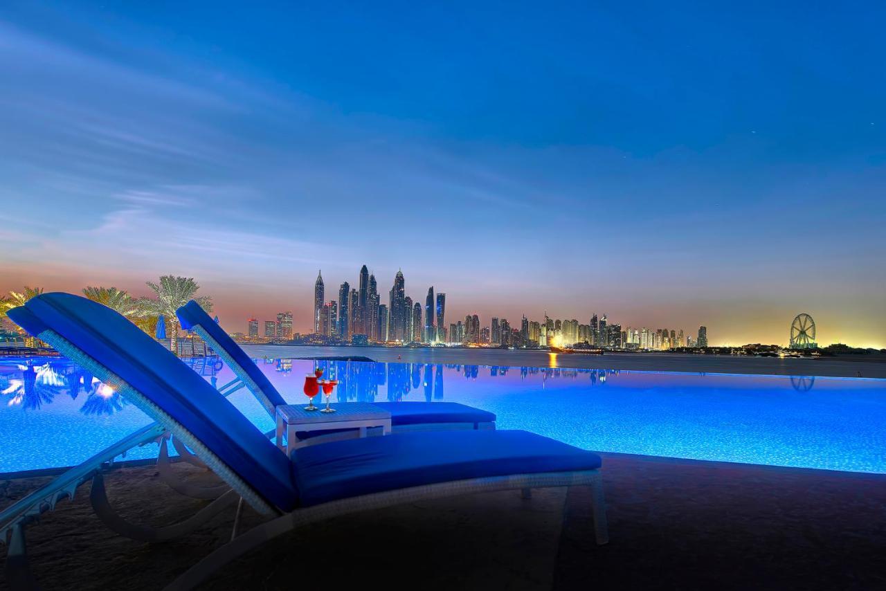 Dukes The Palm, A Royal Hideaway Hotel Dubai Esterno foto The image depicts a tranquil scene by a swimming pool, illuminated by soft blue lighting. In the foreground, there are two lounge chairs positioned beside the pool, suggesting a relaxing environment. A small table between the chairs holds two cocktai