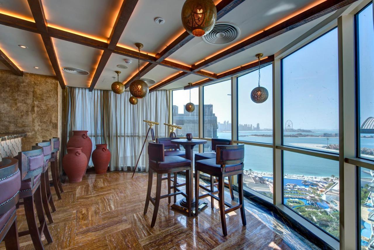Dukes The Palm, A Royal Hideaway Hotel Dubai Esterno foto The photo depicts a modern, stylish interior of a restaurant or lounge. It features large windows that offer a view of a waterfront and cityscape. The design includes warm wooden beams and ambient lighting, creating a cozy atmosphere. There are sever