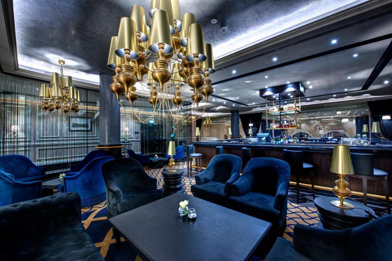Dukes The Palm, A Royal Hideaway Hotel Dubai Esterno foto The photo displays an elegant interior of a lounge or bar area. It features plush blue seating, including armchairs around a dark table. A prominent chandelier with golden lamps hangs from the ceiling, providing ambient light. In the background, ther