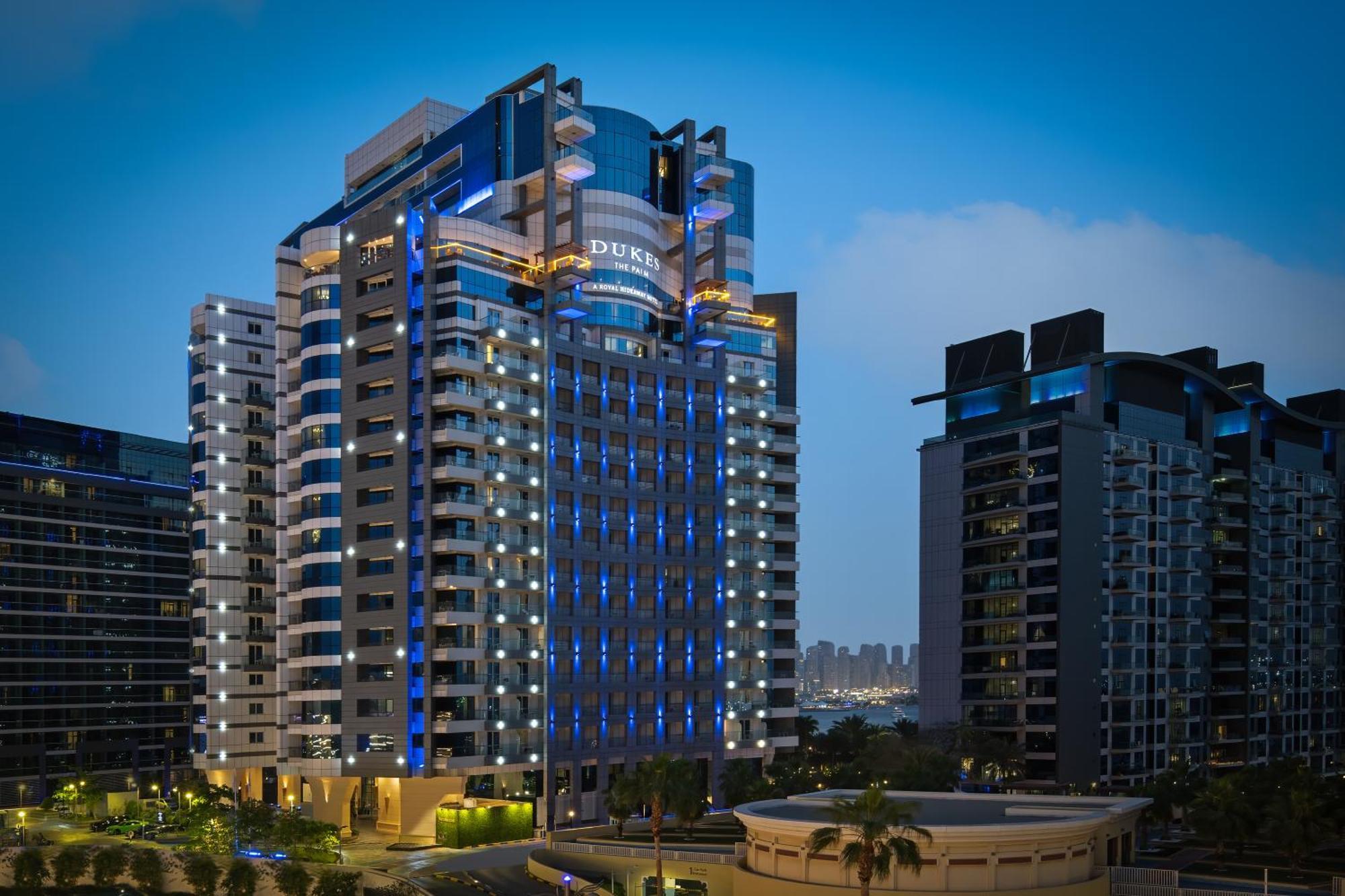 Dukes The Palm, A Royal Hideaway Hotel Dubai Esterno foto The photo shows a modern high-rise building, which appears to be a hotel or residential complex. The design features sleek lines and a glass façade, illuminated with blue lighting that enhances its contemporary look. Surrounding the main building are