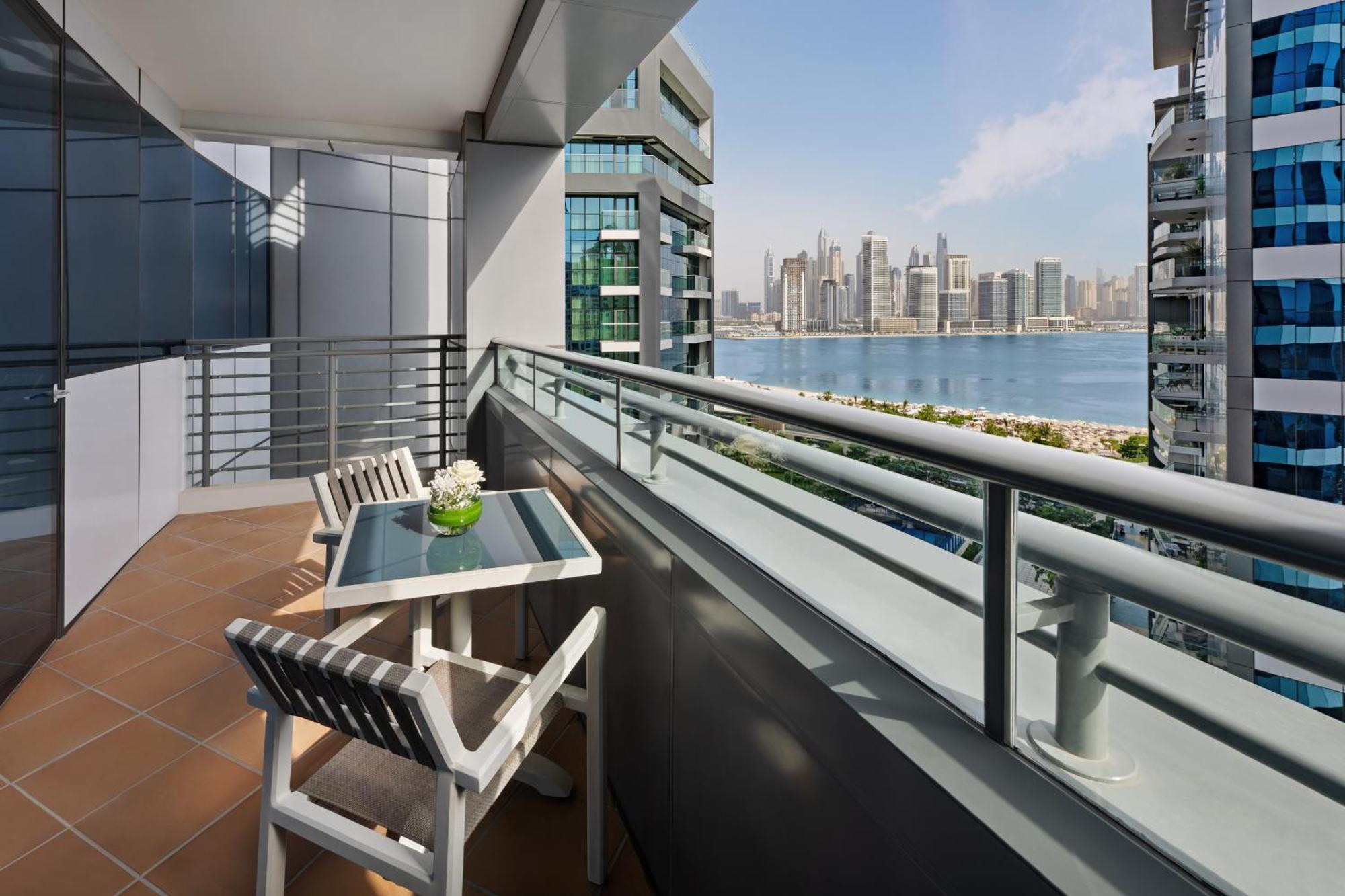 Dukes The Palm, A Royal Hideaway Hotel Dubai Esterno foto The photo shows a balcony with a modern design, featuring a small table and two white chairs. On the table, there is a vase with flowers. The balcony offers a view of a waterfront cityscape, with tall buildings in the background and a calm body of wa