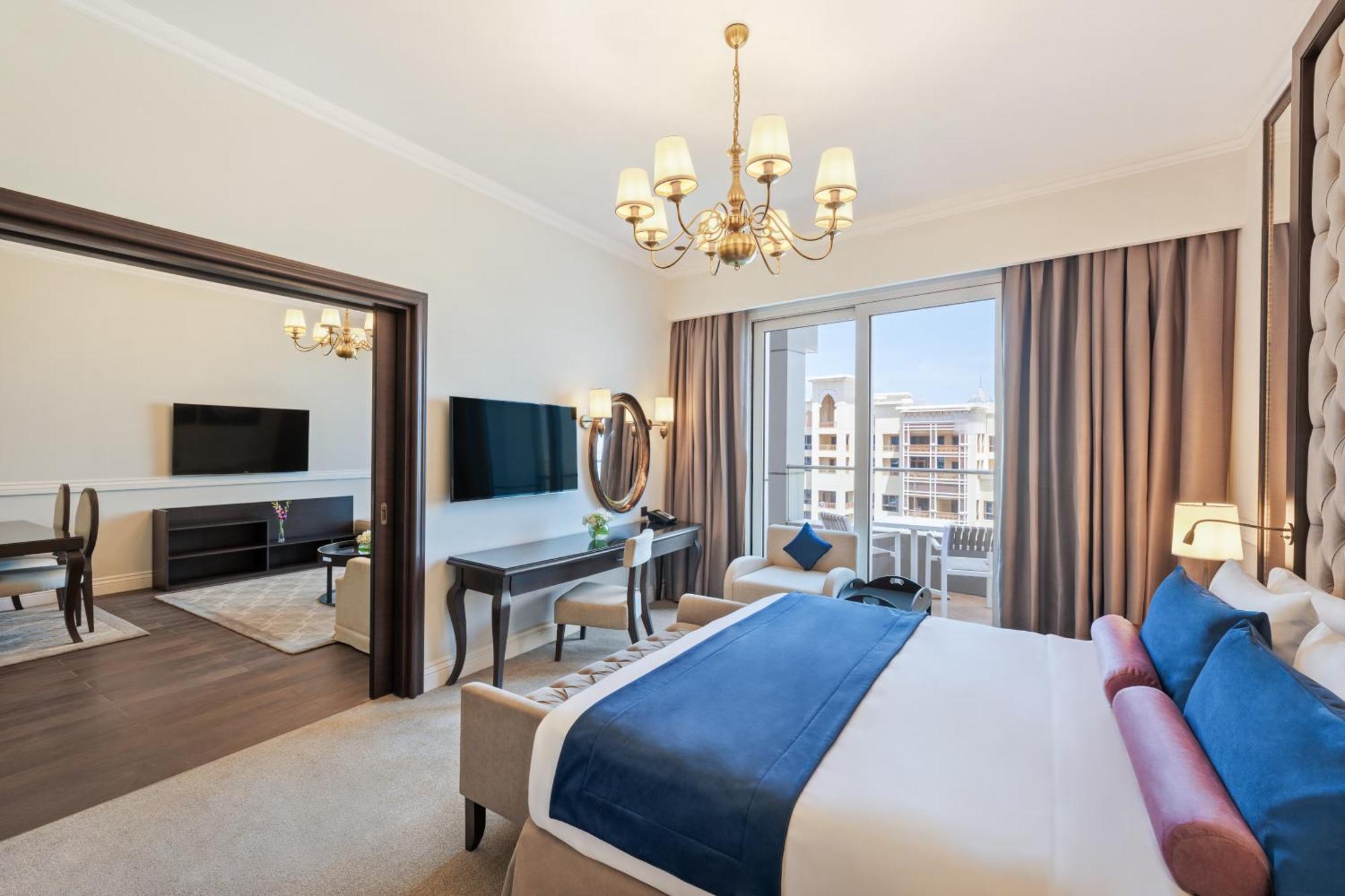 Dukes The Palm, A Royal Hideaway Hotel Dubai Esterno foto The photo depicts a stylish hotel room. It features a comfortable bed with blue and white bedding. The room includes a large window with curtains that allow natural light in, and there are two flat-screen TVs mounted on the walls. A chandelier hangs 
