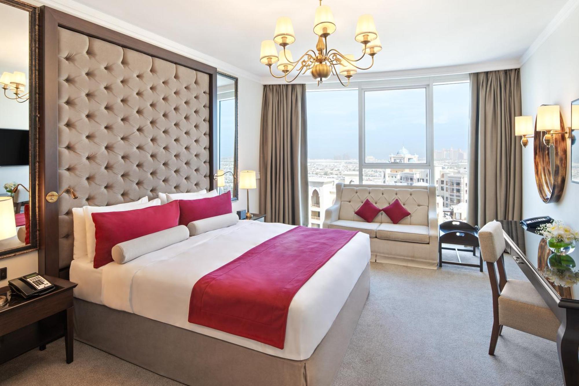Dukes The Palm, A Royal Hideaway Hotel Dubai Esterno foto The photo shows a luxurious hotel room featuring a large, well-made bed with a plush headboard. The bed is adorned with decorative cushions, primarily in shades of red. There is a stylish sofa nearby, also featuring similar color accents. The room is