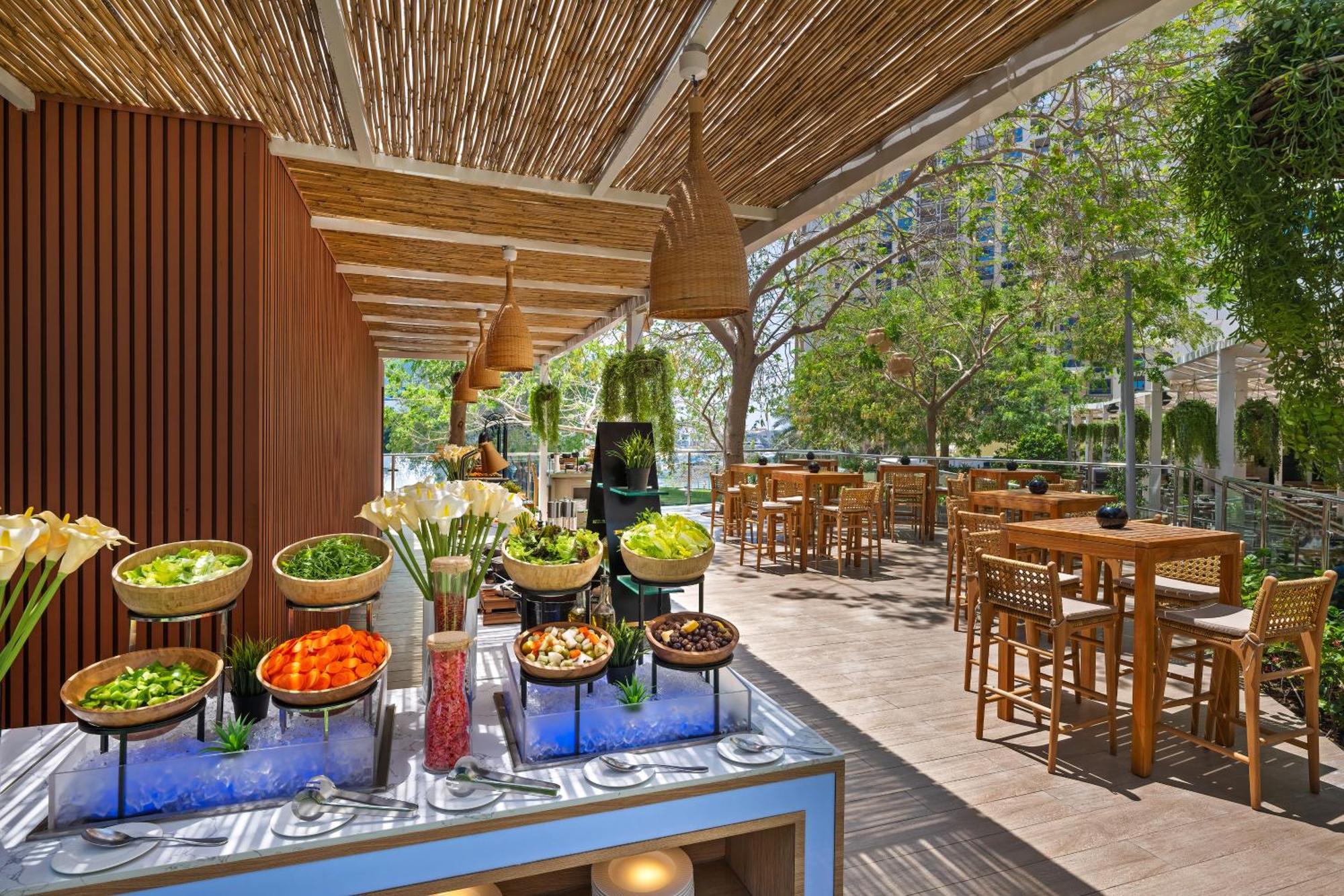 Dukes The Palm, A Royal Hideaway Hotel Dubai Esterno foto The photo showcases a bright and inviting outdoor dining space. In the foreground, there is a buffet table with various salads and fresh vegetables displayed in bowls, including items like lettuce, carrots, and other colorful ingredients, set against