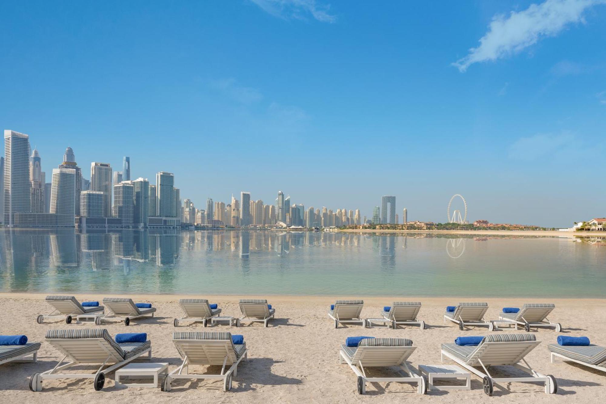 Dukes The Palm, A Royal Hideaway Hotel Dubai Esterno foto The photo shows a serene beach scene with several lounge chairs arranged in the foreground. These chairs are positioned on soft sand and have blue cushions. In the background, there is a stunning view of a modern skyline featuring tall buildings and 