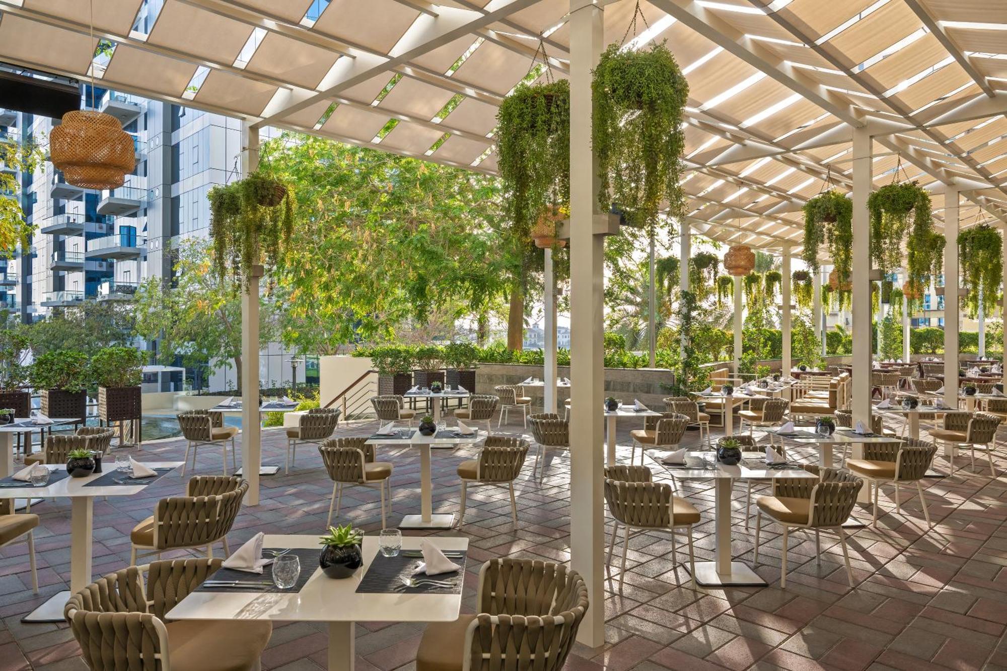 Dukes The Palm, A Royal Hideaway Hotel Dubai Esterno foto The photo shows a charming outdoor restaurant or café with a spacious seating area. The space is adorned with several plants hanging from the ceiling, creating a vibrant and lush atmosphere. There are rows of tables set with utensils and small decora