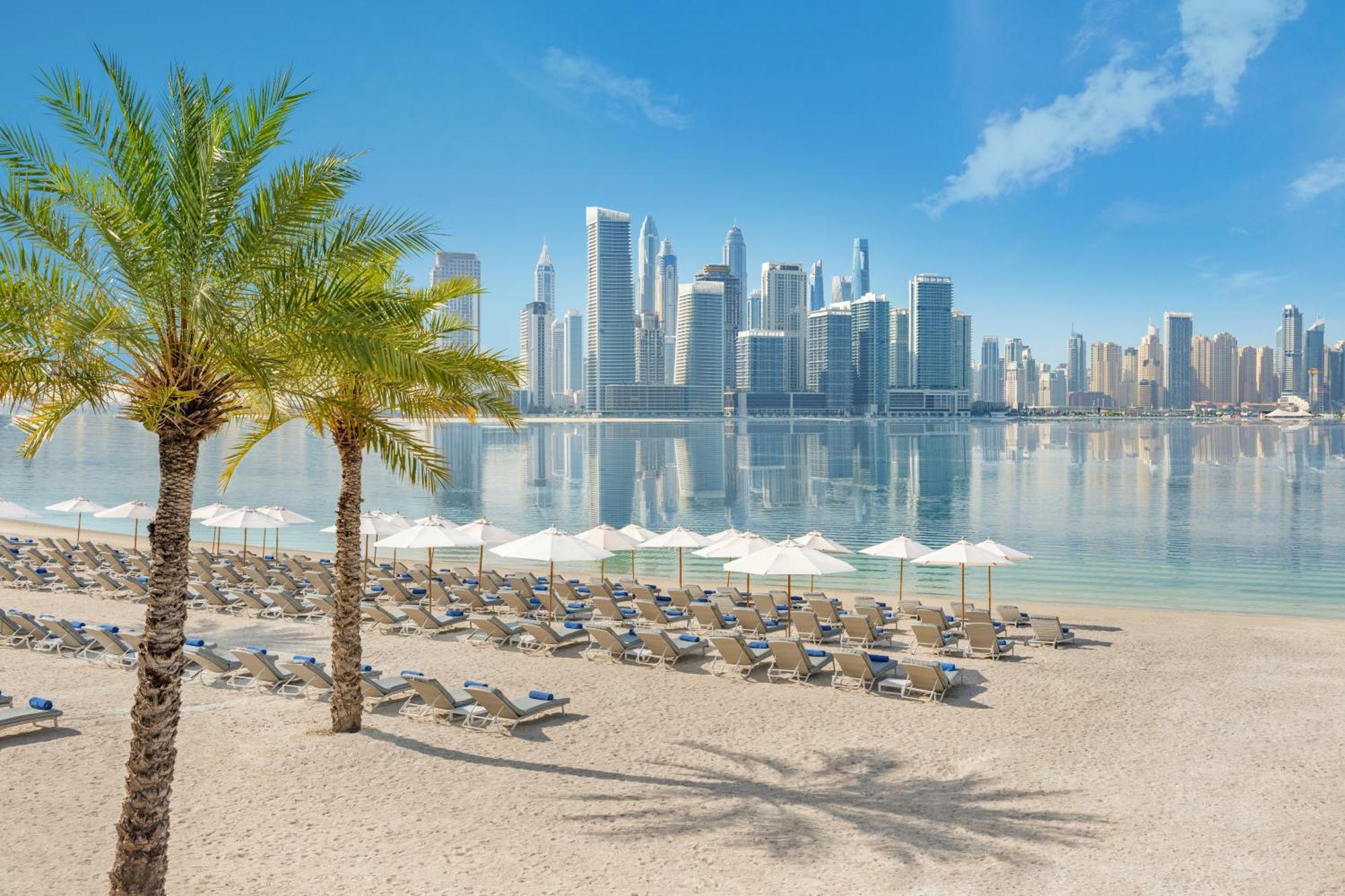 Dukes The Palm, A Royal Hideaway Hotel Dubai Esterno foto The photo depicts a tranquil beach scene with a view of a modern city skyline in the background. In the foreground, there are several lounge chairs arranged neatly on the sandy beach, accompanied by white umbrellas for shade. Palm trees add a tropica