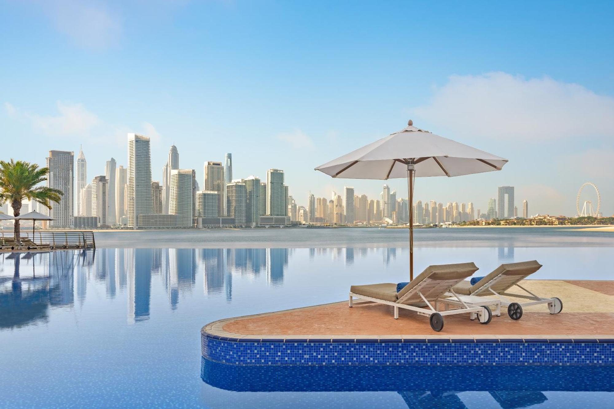 Dukes The Palm, A Royal Hideaway Hotel Dubai Esterno foto The photo shows a serene and luxurious setting, featuring an infinity pool that seems to blend into a body of water. In the foreground, there are two lounge chairs positioned on a platform, accompanied by a large white umbrella providing shade. The b
