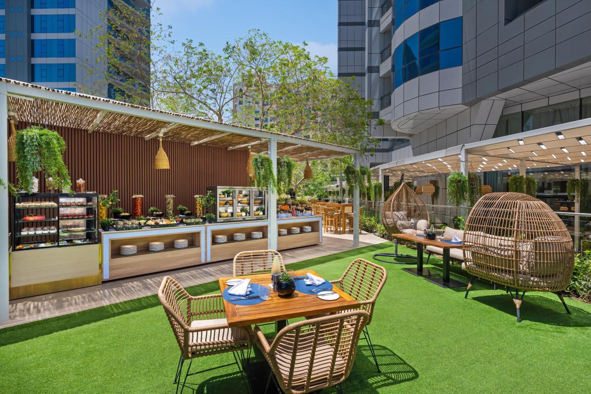 Dukes The Palm, A Royal Hideaway Hotel Dubai Esterno foto The photo shows a modern outdoor dining area, likely part of a restaurant or cafe. There are several seating arrangements including a wooden table surrounded by four wicker chairs. On the table, there are blue plates and a decorative item in the cent