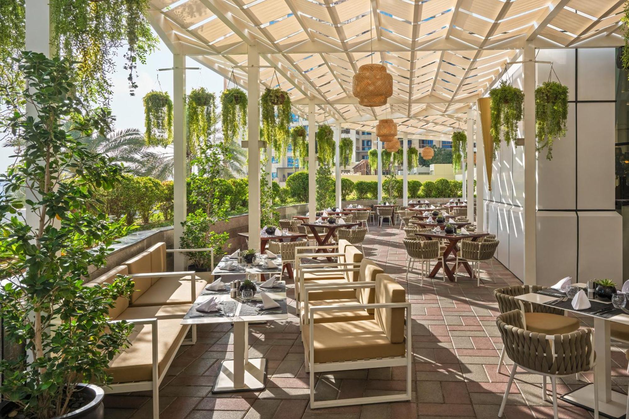 Dukes The Palm, A Royal Hideaway Hotel Dubai Esterno foto The photo shows an outdoor restaurant patio area with a bright and inviting atmosphere. There are several tables and chairs arranged neatly under a structured canopy. The furniture appears to be a mix of light-colored wooden pieces and wicker chairs.