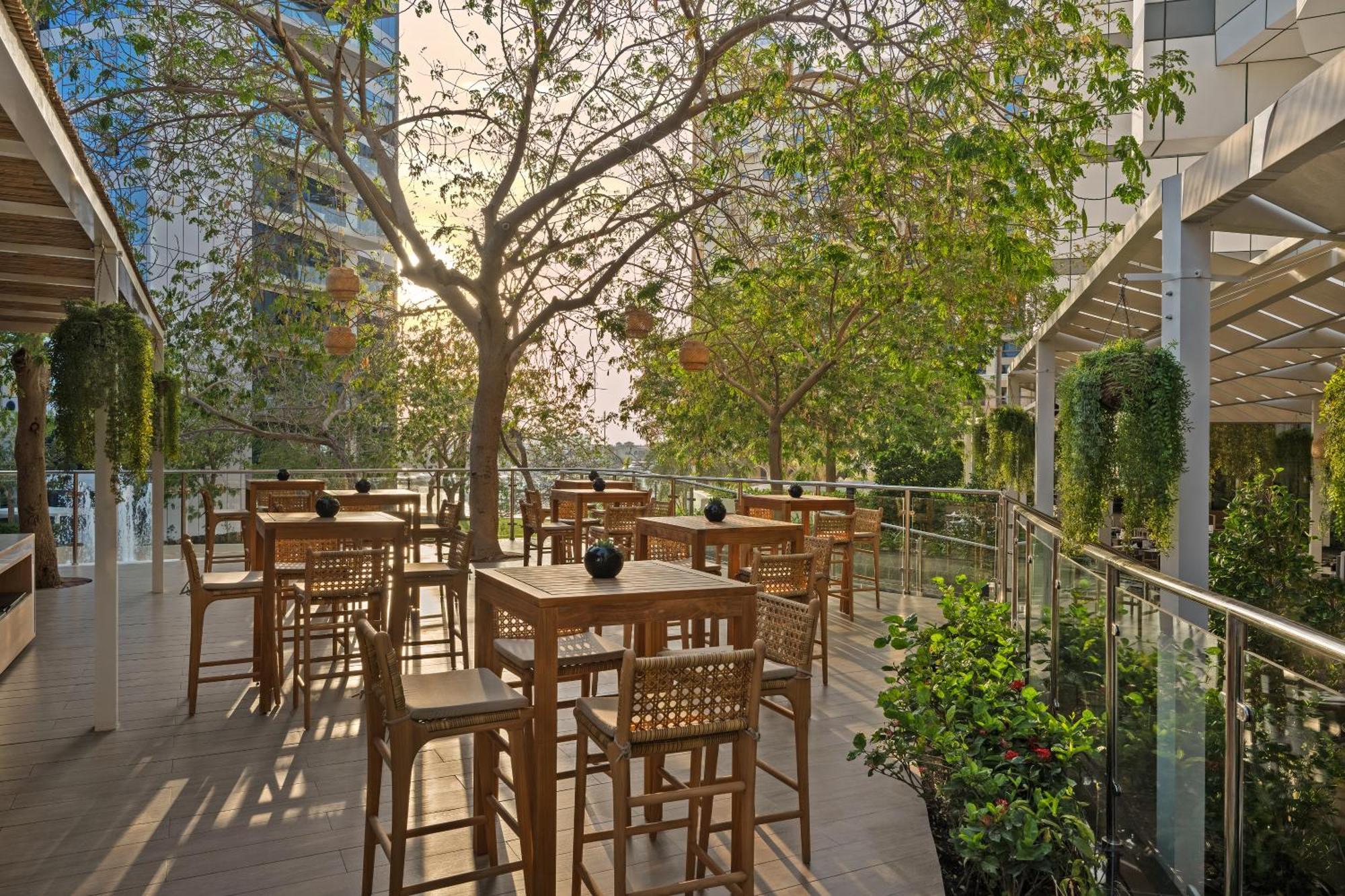 Dukes The Palm, A Royal Hideaway Hotel Dubai Esterno foto The photo shows a spacious outdoor dining area featuring wooden tables and chairs. There is a large tree with green leaves that provides shade, and the ambiance is enhanced by natural light filtering through the leaves. Surrounding the area are glass