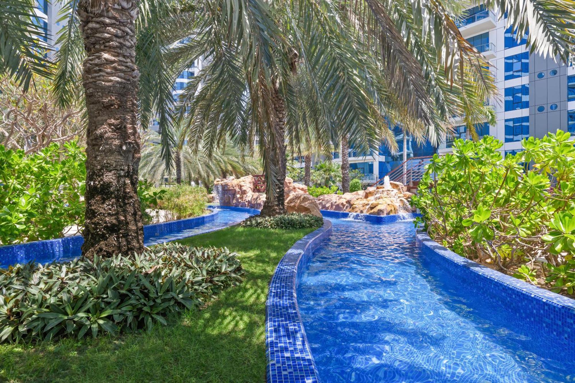 Dukes The Palm, A Royal Hideaway Hotel Dubai Esterno foto The photo depicts a beautifully landscaped tropical area featuring a winding waterway with clear blue water, bordered by lush greenery and palm trees. The vibrant blue tiles lining the water add a decorative touch, while the overall setting suggests 