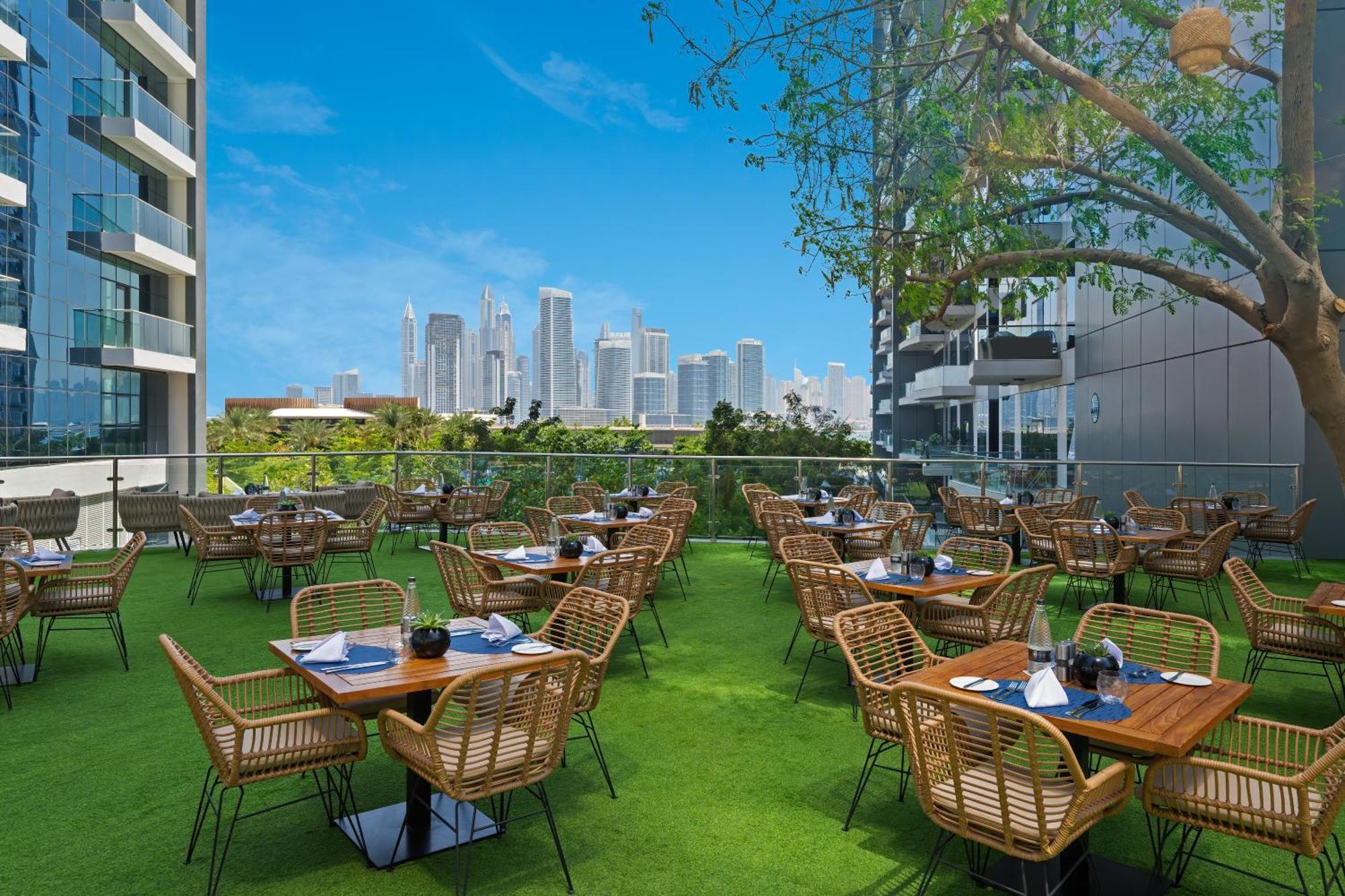 Dukes The Palm, A Royal Hideaway Hotel Dubai Esterno foto The photo shows an outdoor dining area with several wooden tables and wicker chairs arranged neatly. The tables are set with napkins and utensils, indicating readiness for guests. The dining area is surrounded by a green lawn and features a tree, pro