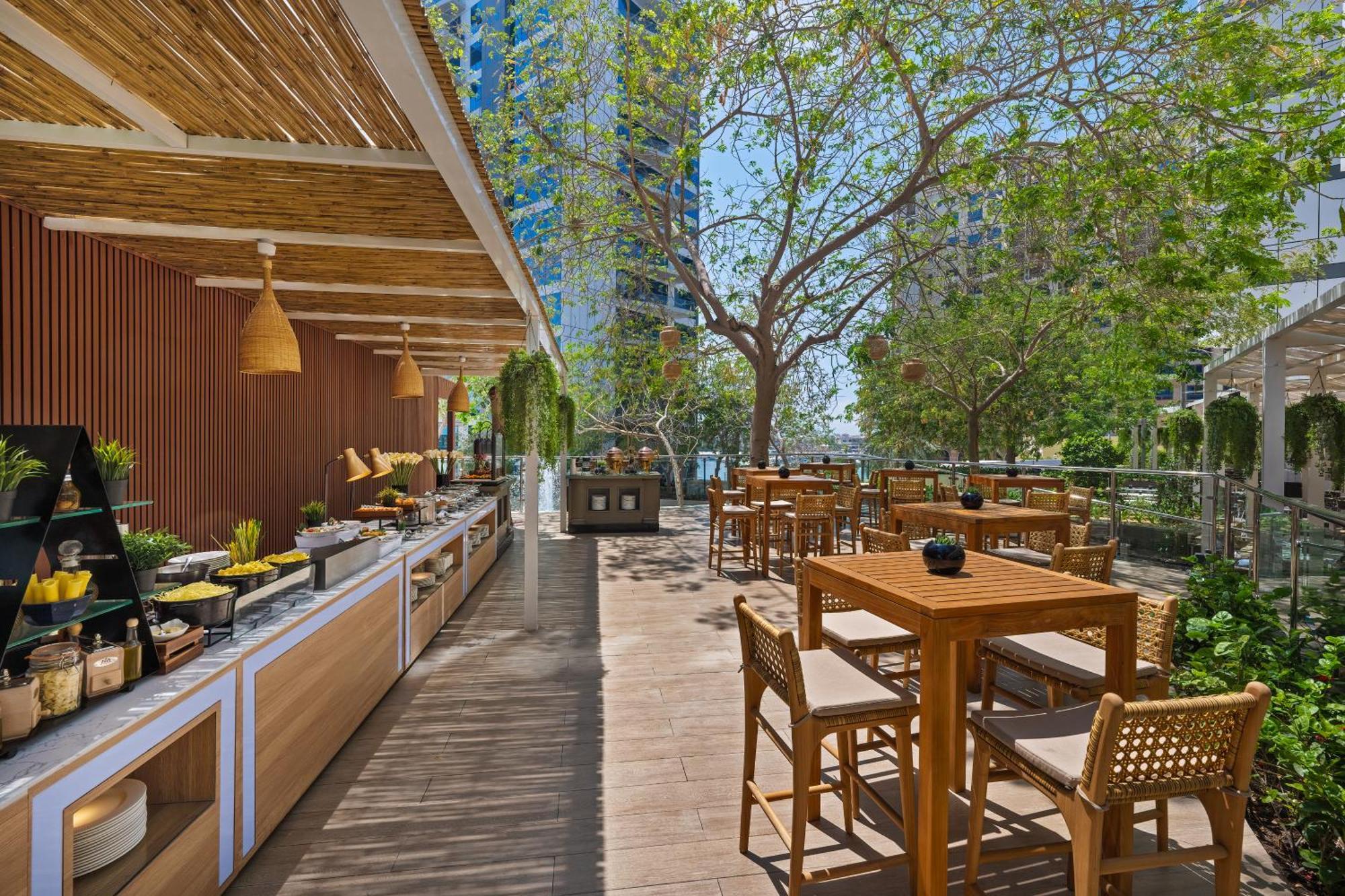 Dukes The Palm, A Royal Hideaway Hotel Dubai Esterno foto The photo showcases an outdoor dining area in a restaurant or café. It features a wooden terrace with several tables and chairs arranged for patrons. On one side, there is a buffet or serving area with various displays of food items. 

Surrounding th