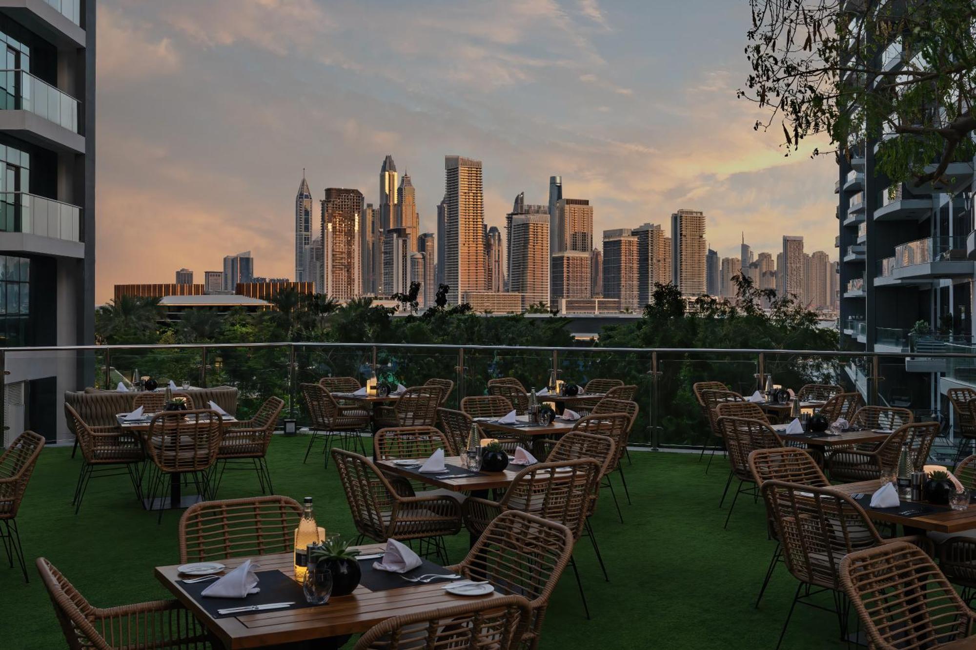 Dukes The Palm, A Royal Hideaway Hotel Dubai Esterno foto The photo shows an outdoor dining area with several tables and chairs arranged neatly. The tables are set with plates and candles, creating an inviting atmosphere. In the background, there is a stunning city skyline featuring tall skyscrapers, bathed
