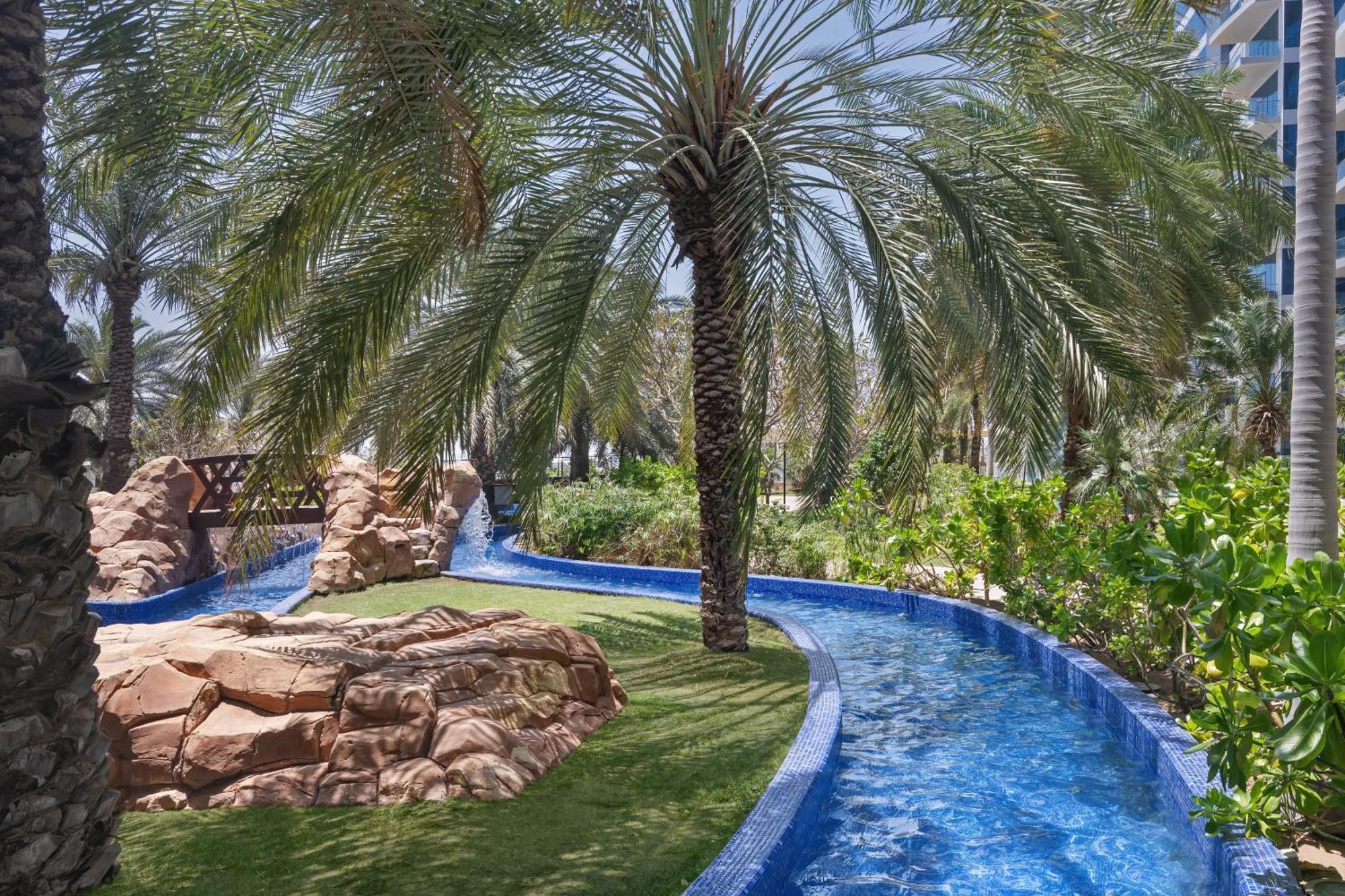 Dukes The Palm, A Royal Hideaway Hotel Dubai Esterno foto The photo depicts a serene outdoor setting, featuring a winding blue water channel surrounded by lush greenery. Palm trees tower over the scene, providing shade and a tropical feel. There are rocky formations integrated into the landscape, and the ov