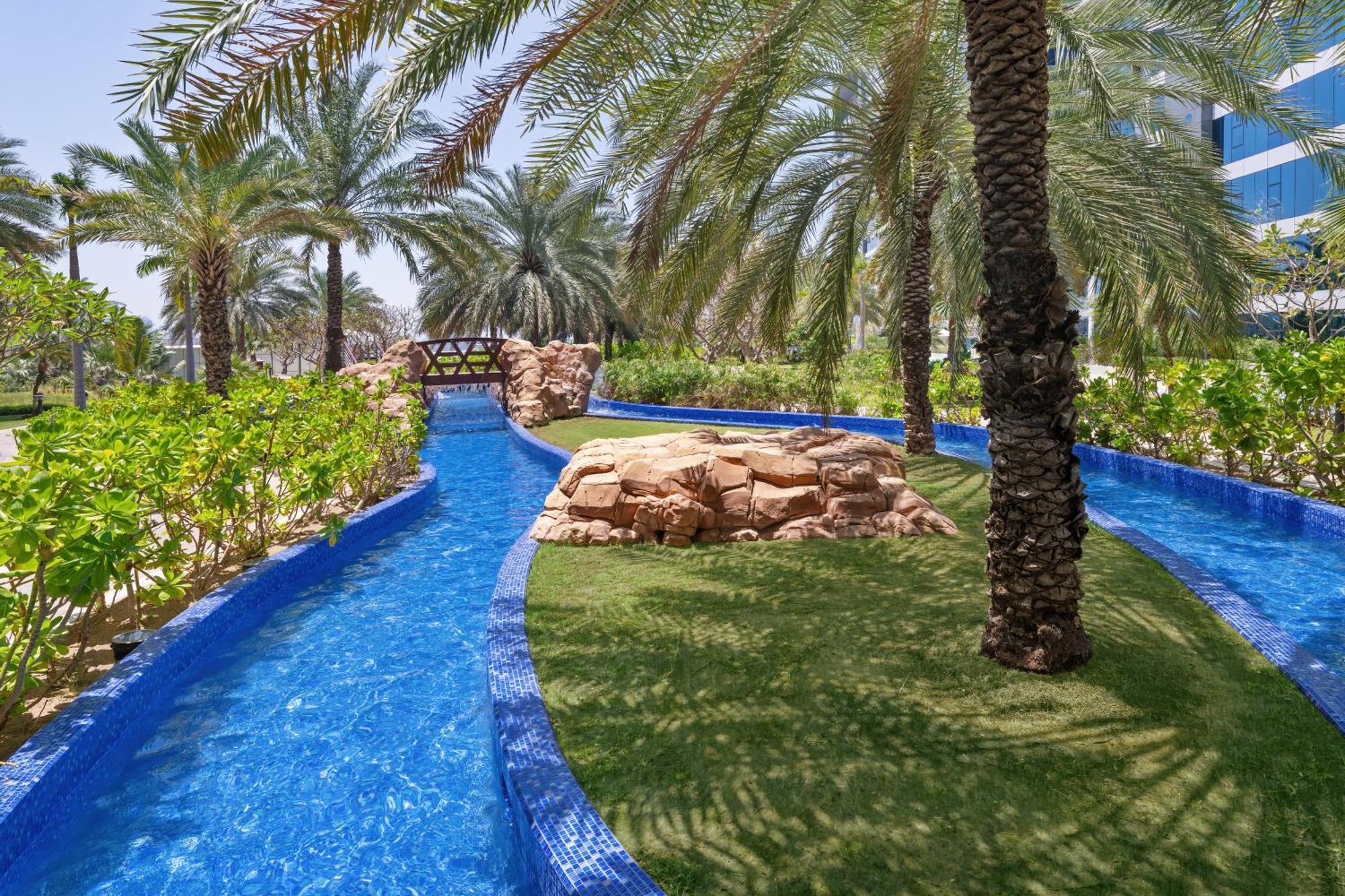 Dukes The Palm, A Royal Hideaway Hotel Dubai Esterno foto The photo depicts a serene outdoor area featuring a winding, clear blue waterway surrounded by lush greenery and palm trees. In the background, there is a small stone bridge arching over the water. Natural rock formations and manicured grass create a