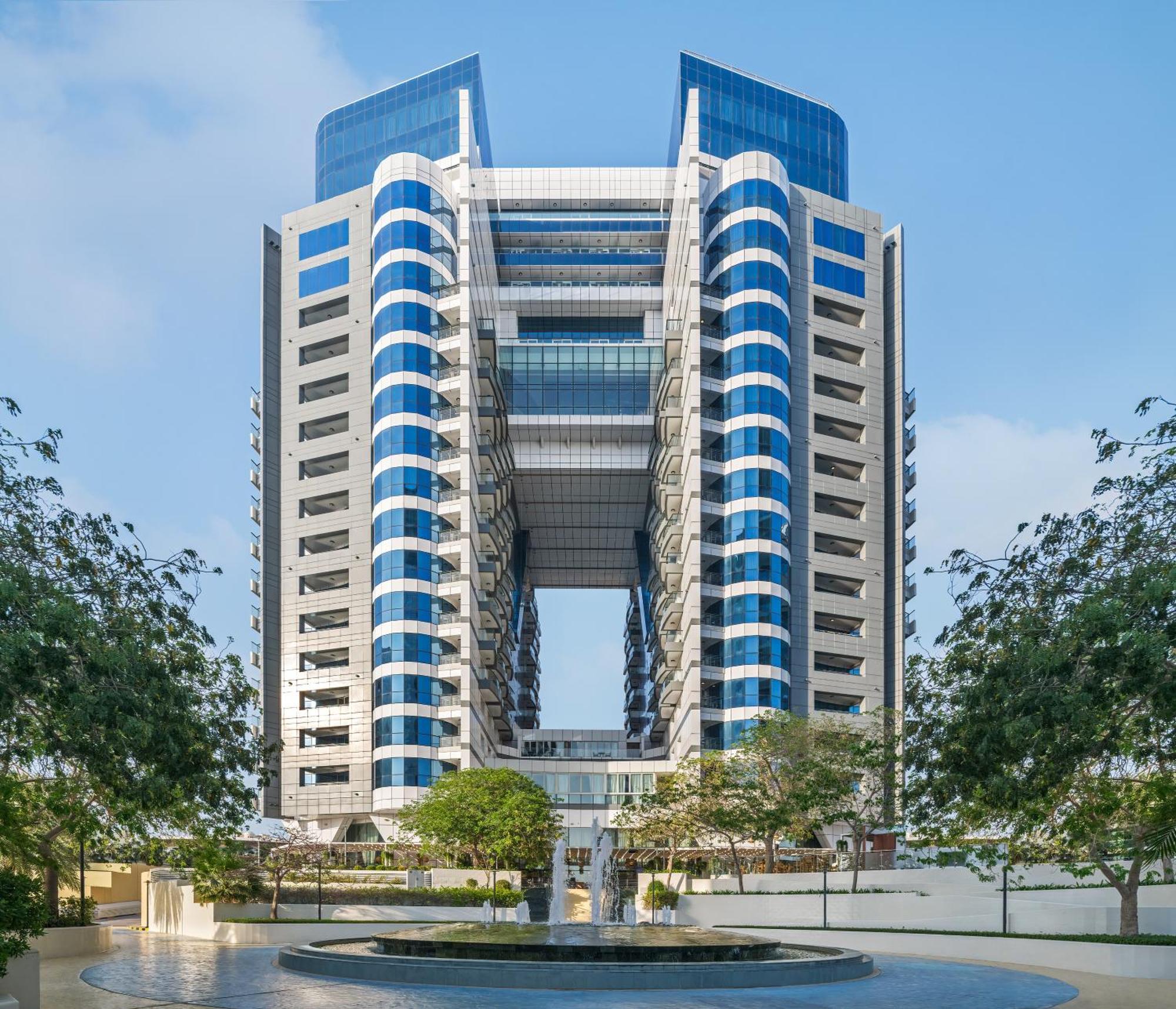 Dukes The Palm, A Royal Hideaway Hotel Dubai Esterno foto The image shows a large modern building characterized by a distinctive architectural design. It features a symmetrical façade with multiple levels and large, blue-tinted glass windows. The structure has prominent vertical elements and an open center,