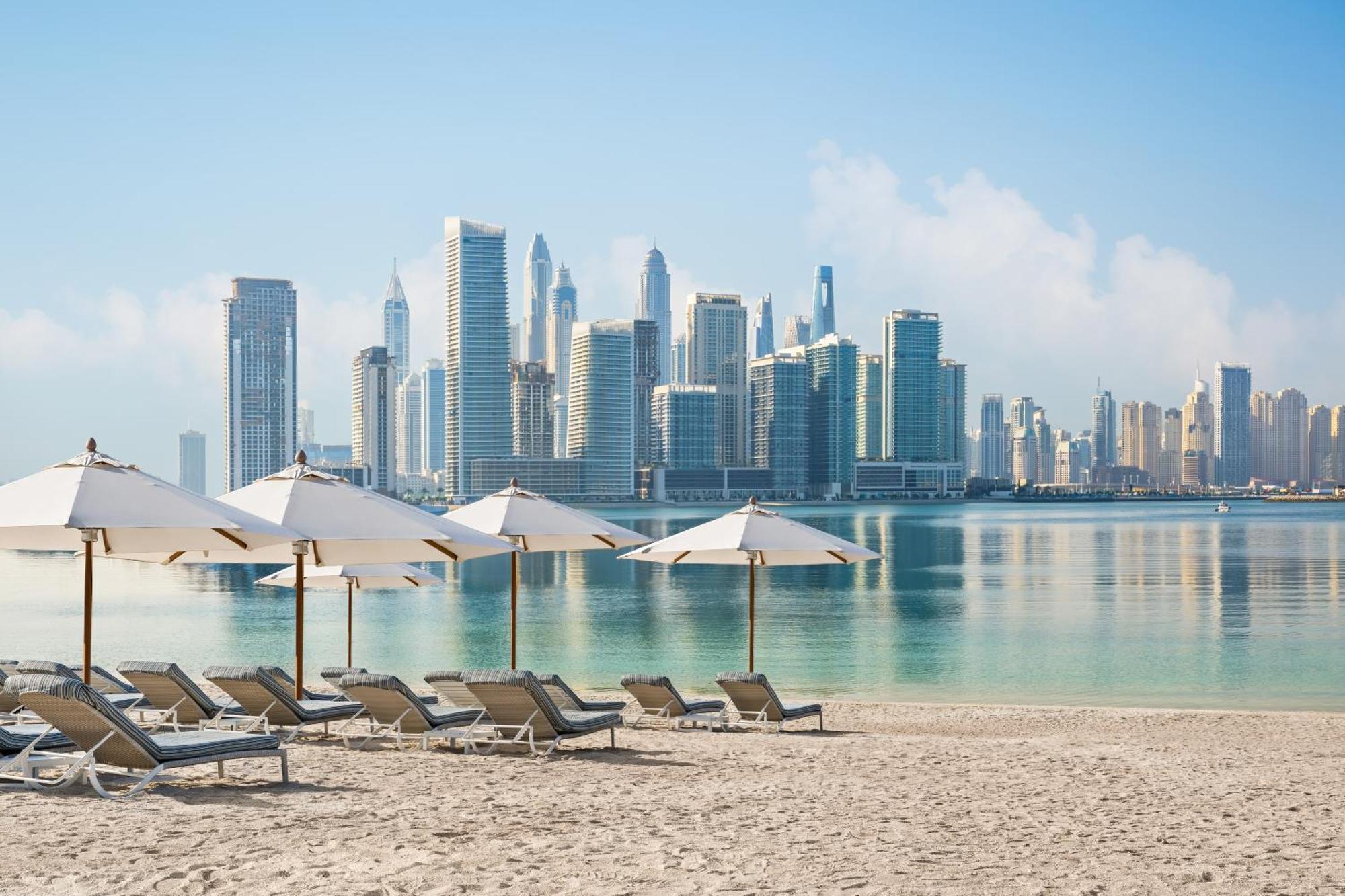 Dukes The Palm, A Royal Hideaway Hotel Dubai Esterno foto The photo showcases a serene beach scene with several lounge chairs positioned on the sandy shore. Each chair is paired with a large umbrella for shade. In the background, a modern city skyline is visible, featuring tall skyscrapers and a clear blue 