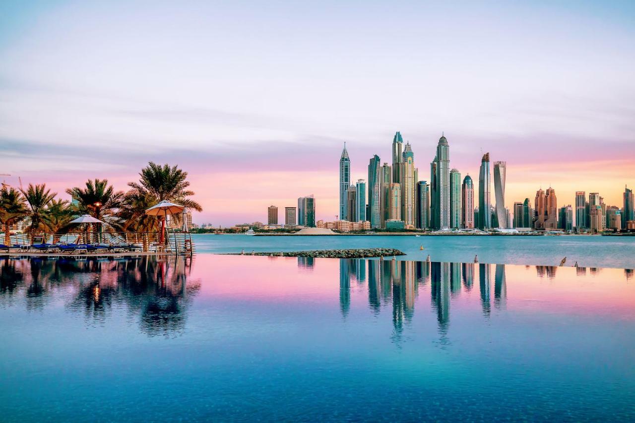 Dukes The Palm, A Royal Hideaway Hotel Dubai Esterno foto The image showcases a stunning skyline view of a modern city, likely during sunrise or sunset, as indicated by the beautiful pastel colors in the sky. The foreground features a calm body of water, reflecting the skyscrapers and the colorful sky. Lush